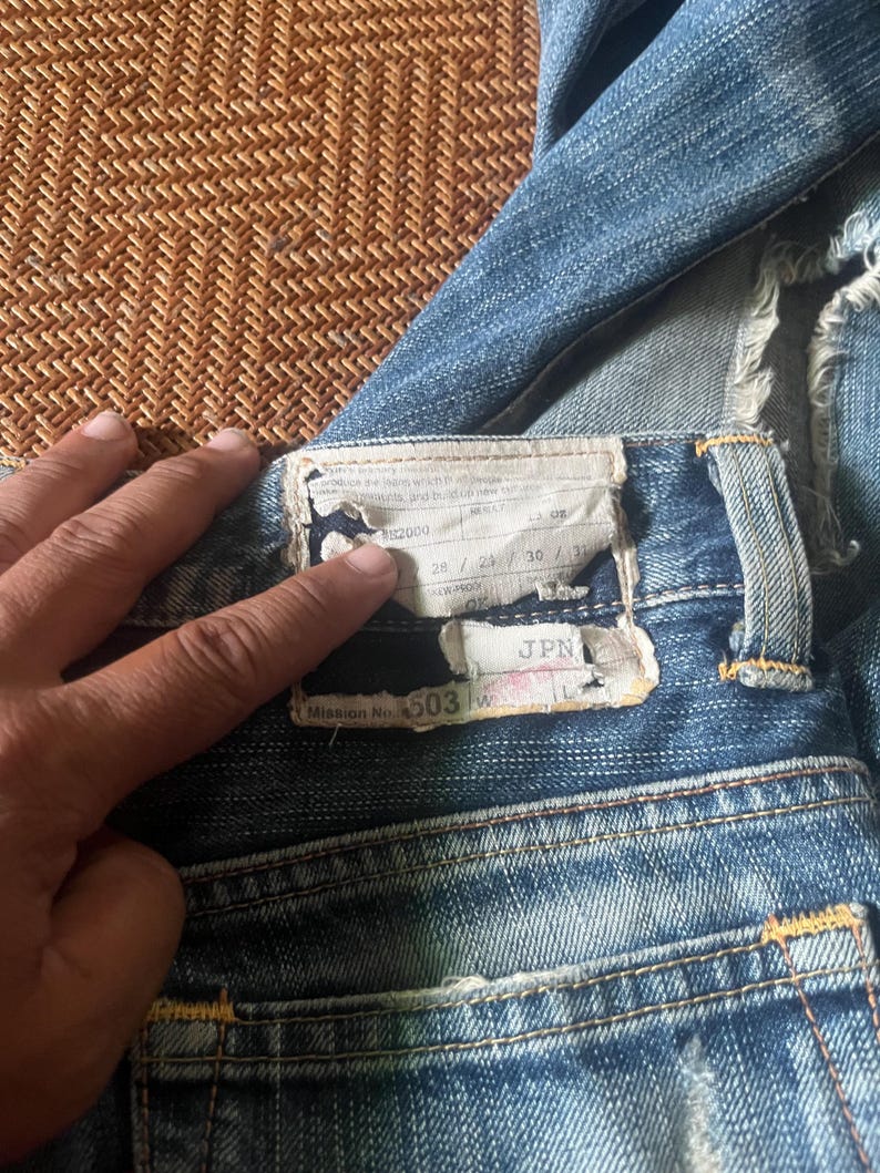 May include: Close-up of blue denim jeans with a worn appearance. The back pocket area is visible, with a label showing size measurements and the text "Mission No. 503". The jeans have a classic five-pocket design and yellow stitching.