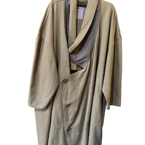 May include: A tan-coloured, oversized coat with an asymmetrical design. The coat has a draped collar, three buttons, and a relaxed fit. It hangs on a black hanger against a white background, highlighting its flowing silhouette.
