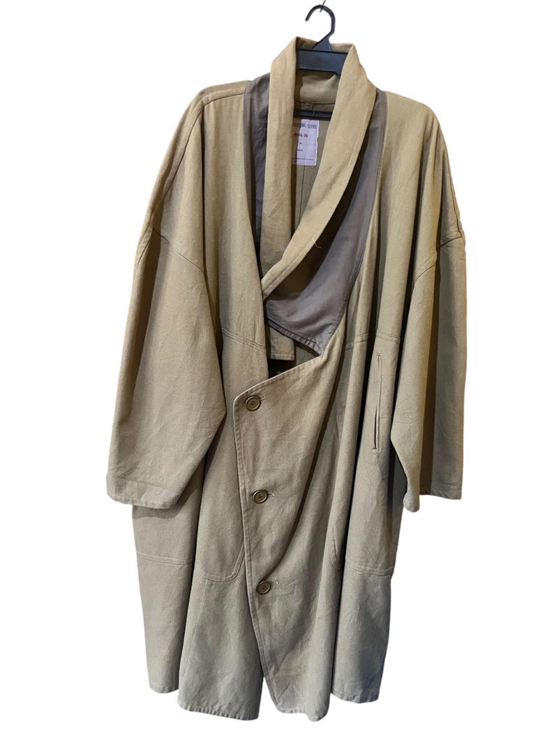 May include: A tan-coloured, oversized coat with an asymmetrical design. The coat has a draped collar, three buttons, and a relaxed fit. It hangs on a black hanger against a white background, highlighting its flowing silhouette.