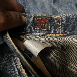 May include: Close-up of blue denim jeans with a red and black Edwin label. The label reads "EDWIN No. 505 SX DESIGN PATENTED". A white tag shows the size 30 x 33. The jeans are slightly faded.