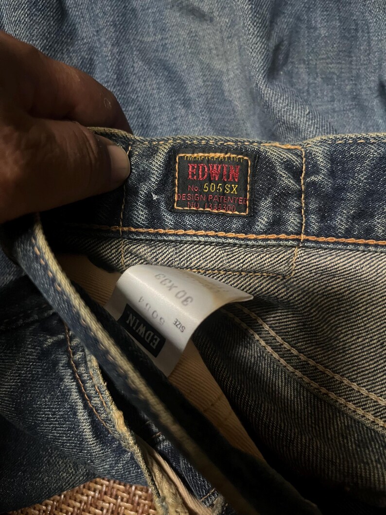May include: Close-up of blue denim jeans with a red and black Edwin label. The label reads "EDWIN No. 505 SX DESIGN PATENTED". A white tag shows the size 30 x 33. The jeans are slightly faded.