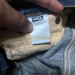 May include: Close-up of a pair of blue denim jeans. The tag reads "EDWIN" with the numbers "5054" and "30x33". The jeans are folded, showing the inside lining and a button.