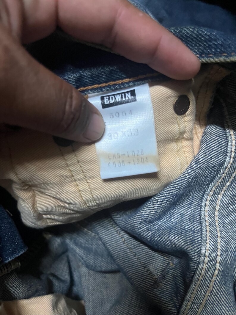 May include: Close-up of a pair of blue denim jeans. The tag reads "EDWIN" with the numbers "5054" and "30x33". The jeans are folded, showing the inside lining and a button.