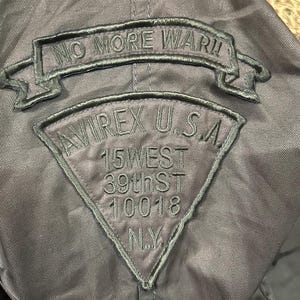 Pu&ograve; includere: Giacca grigio scuro con toppe ricamate. La toppa superiore recita "NO MORE WAR!!" in un banner. Sotto, una toppa triangolare dice "AVIREX U.S.A." con l'indirizzo "15 WEST 39th ST 10018 N.Y."
