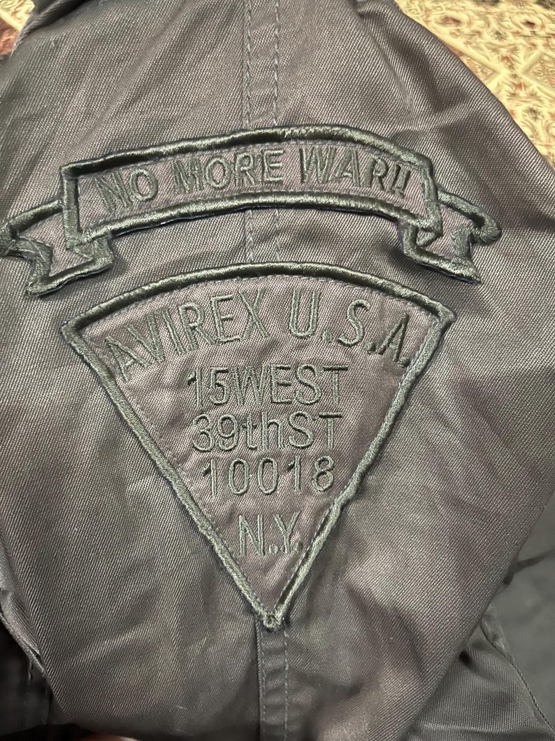 Pu&ograve; includere: Giacca grigio scuro con toppe ricamate. La toppa superiore recita "NO MORE WAR!!" in un banner. Sotto, una toppa triangolare dice "AVIREX U.S.A." con l'indirizzo "15 WEST 39th ST 10018 N.Y."