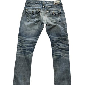 May include: Faded blue denim jeans with a classic five-pocket design. Features decorative stitching on the back pockets and a leather patch at the waistband. The jeans have a worn, distressed look.