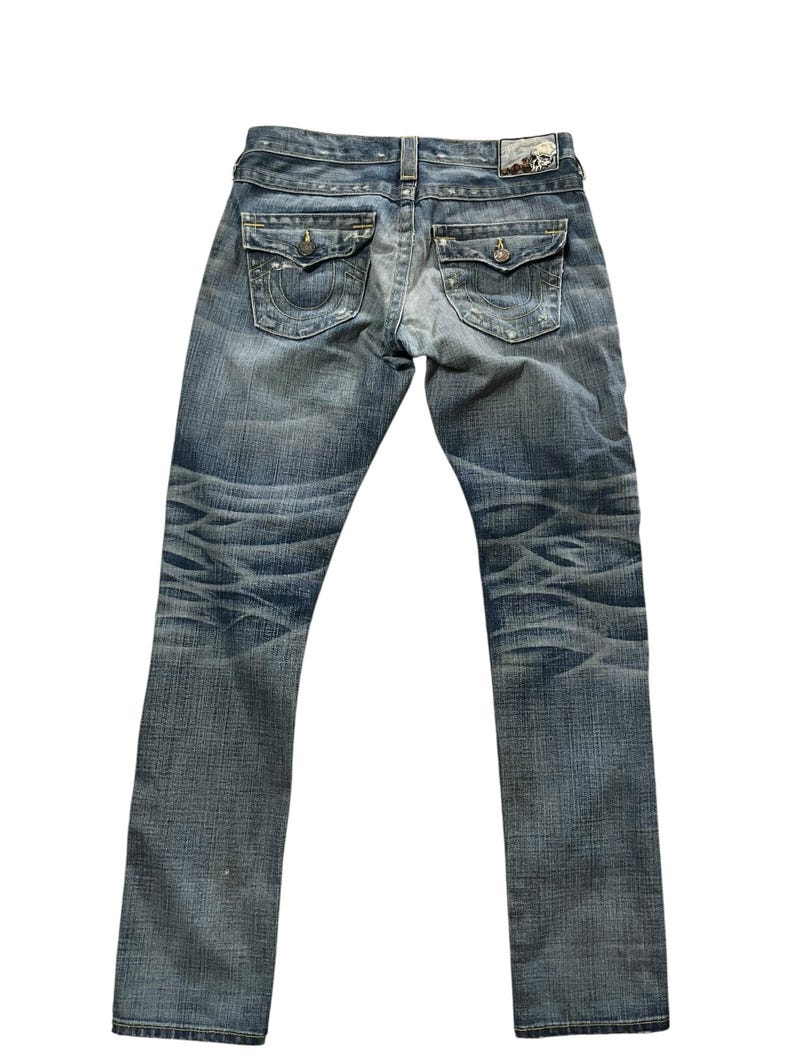 May include: Faded blue denim jeans with a classic five-pocket design. Features decorative stitching on the back pockets and a leather patch at the waistband. The jeans have a worn, distressed look.