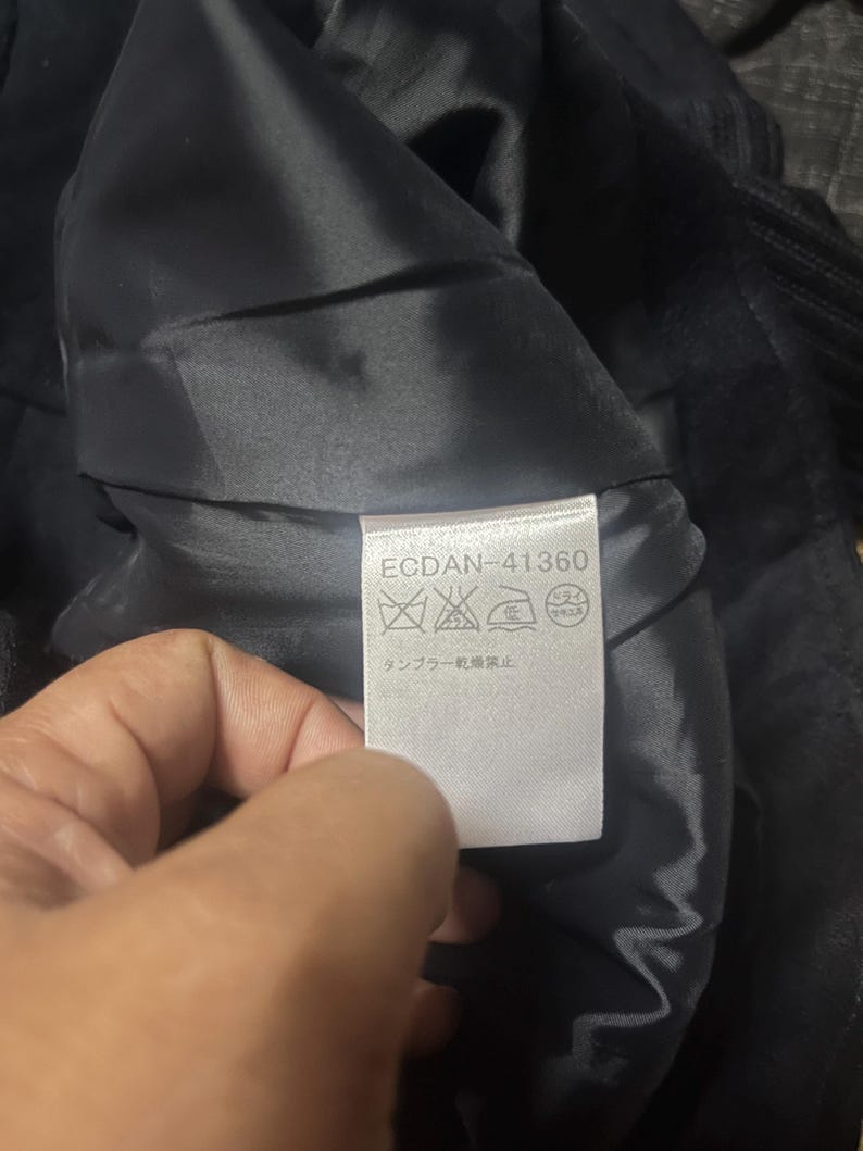 May include: A close-up of a black garment's interior, revealing a tag with the text "ECDAN-41360" and care symbols. The fabric appears to be a dark, possibly velvet or satin material.