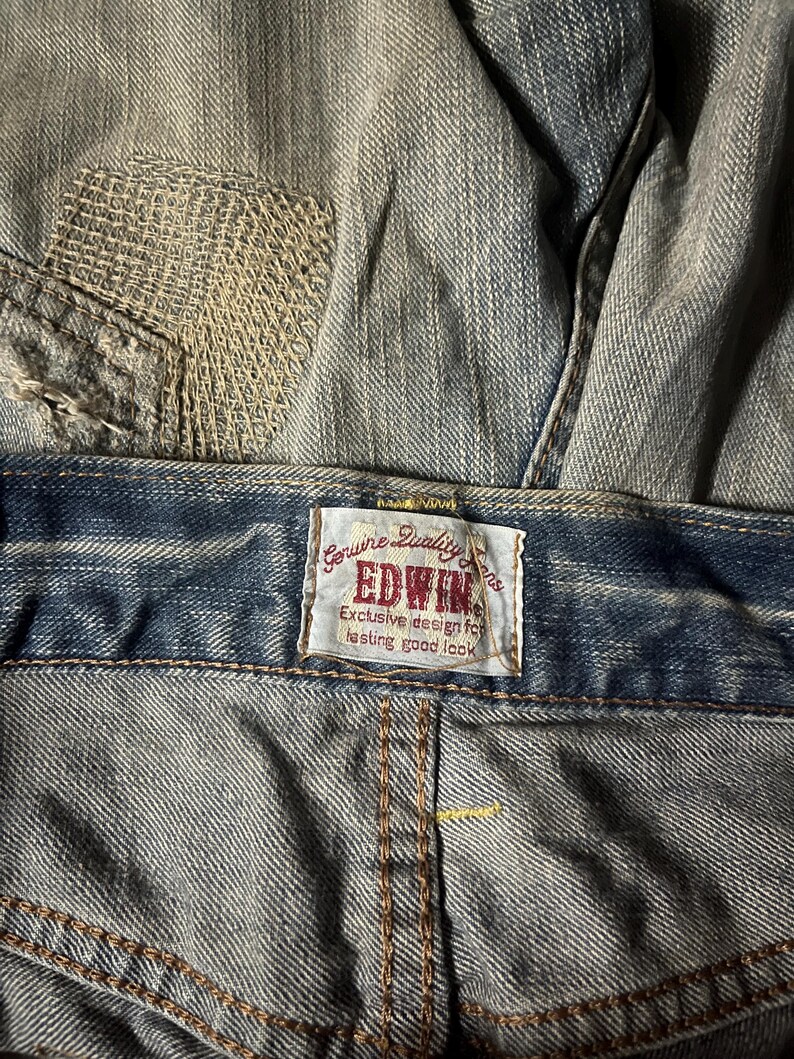 K&ouml;nnte beinhalten: Nahaufnahme einer blauen Jeans mit einem sichtbaren Patch und dem Markenetikett "EDWIN". Auf dem Etikett steht "Genuine Quality Jeans" und "Exclusive design for lasting good look".