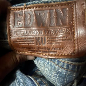 K&ouml;nnte beinhalten: Nahaufnahme eines braunen Lederpatches auf einer blauen Jeans. Der Patch ist mit dem Markennamen "EDWIN" und dem Text "EXCLUSIVE VINTAGE" gepr&auml;gt. Die Jeans hat sichtbare N&auml;hte und eine verwaschene Optik.
