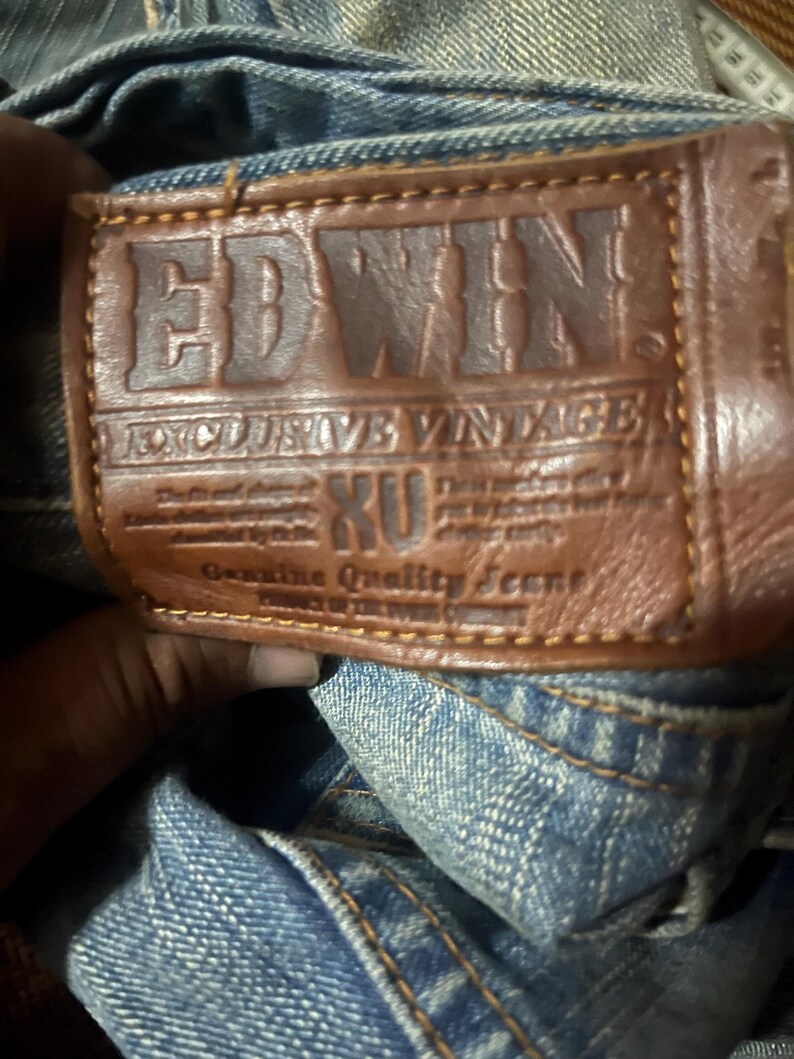K&ouml;nnte beinhalten: Nahaufnahme eines braunen Lederpatches auf einer blauen Jeans. Der Patch ist mit dem Markennamen "EDWIN" und dem Text "EXCLUSIVE VINTAGE" gepr&auml;gt. Die Jeans hat sichtbare N&auml;hte und eine verwaschene Optik.