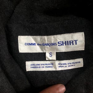 May include: Close-up of a black garment tag with the text "COMME des GAR&Ccedil;ONS SHIRT" in white. Below the text is a white tag with the letter "S". Two additional tags display fabric composition and "FABRIQUE EN FRANCE" and "MADE IN FRANCE".