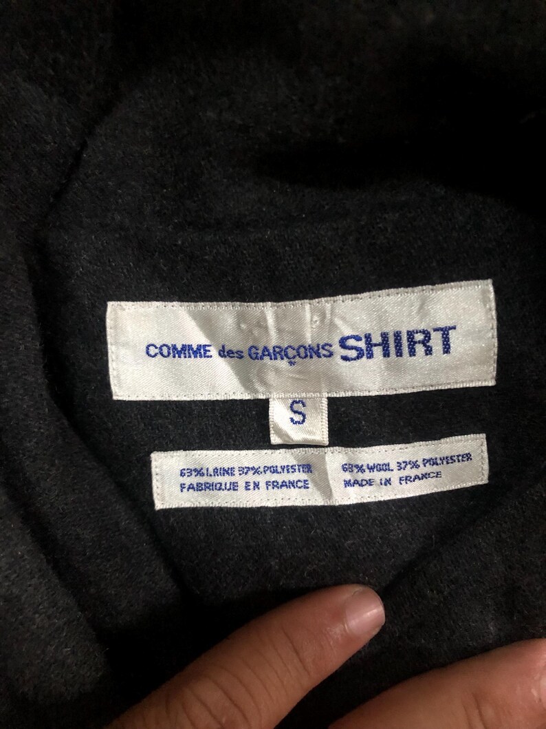 May include: Close-up of a black garment tag with the text "COMME des GAR&Ccedil;ONS SHIRT" in white. Below the text is a white tag with the letter "S". Two additional tags display fabric composition and "FABRIQUE EN FRANCE" and "MADE IN FRANCE".