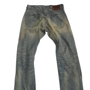 K&ouml;nnte beinhalten: Verblasste blaue Jeans mit einem Used-Look. Die Jeans haben ein klassisches F&uuml;nf-Taschen-Design, einen braunen Lederaufn&auml;her und ein rotes Etikett auf der Ges&auml;&szlig;tasche. Die Jeans weisen Gebrauchsspuren auf.