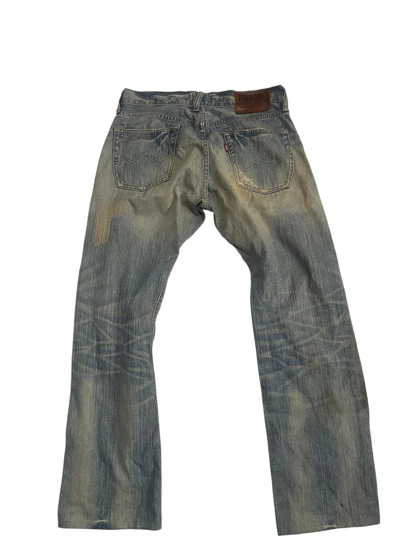 K&ouml;nnte beinhalten: Verblasste blaue Jeans mit einem Used-Look. Die Jeans haben ein klassisches F&uuml;nf-Taschen-Design, einen braunen Lederaufn&auml;her und ein rotes Etikett auf der Ges&auml;&szlig;tasche. Die Jeans weisen Gebrauchsspuren auf.