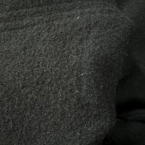 May include: Close-up of a dark gray fabric with a soft, textured surface. The material appears to be a thick knit or fleece, with visible fibers and a slightly uneven texture. The fabric is folded, creating shadows and depth.