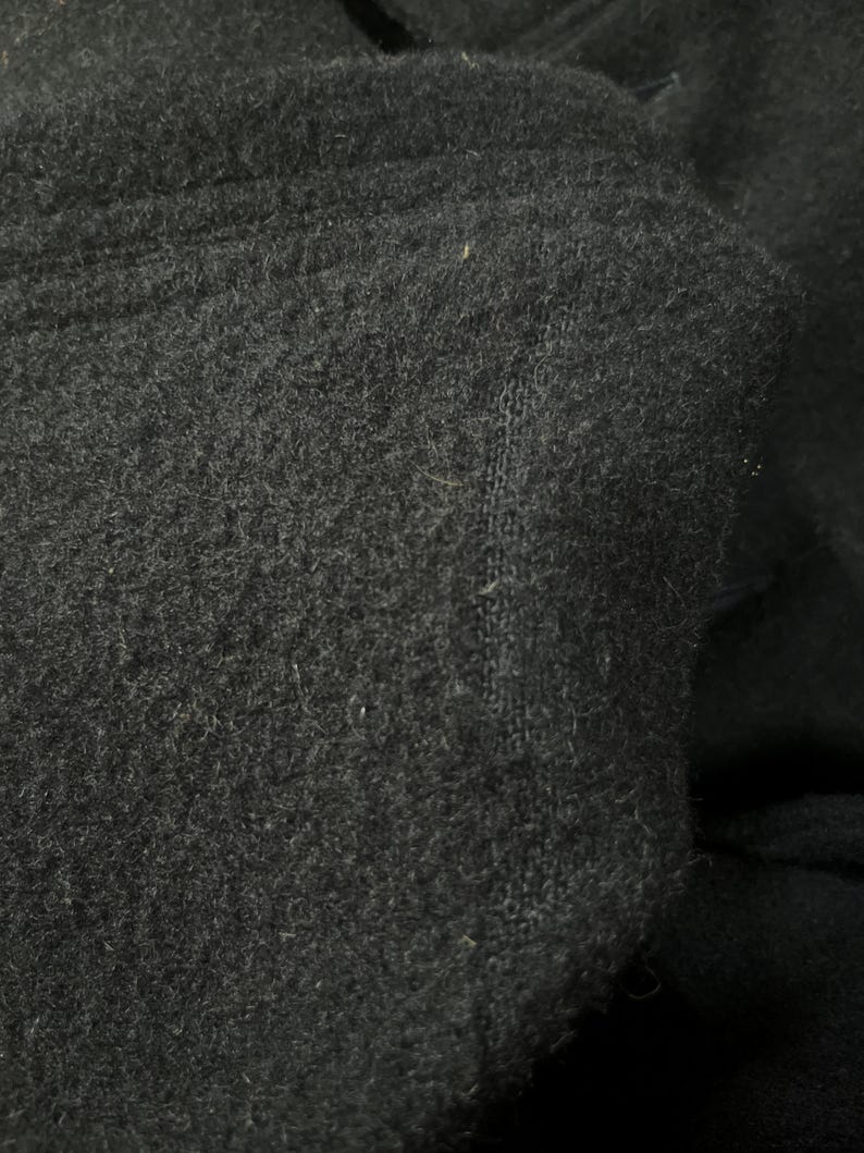 May include: Close-up of a dark gray fabric with a soft, textured surface. The material appears to be a thick knit or fleece, with visible fibers and a slightly uneven texture. The fabric is folded, creating shadows and depth.