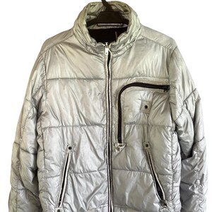 May include: A metallic silver puffer jacket featuring a high collar, a full-length front zipper, and ribbed knit cuffs and hem. The jacket includes two lower zippered pockets and one asymmetrical zippered chest pocket with black trim details.