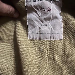 May include: A close-up of a clothing label. The label reads "OLD TRADITIONAL CLOTHES" at the top, with "Moldy fig" below. The label also includes "by Y's bis" and a Tokyo address. The garment is a light olive green colour.