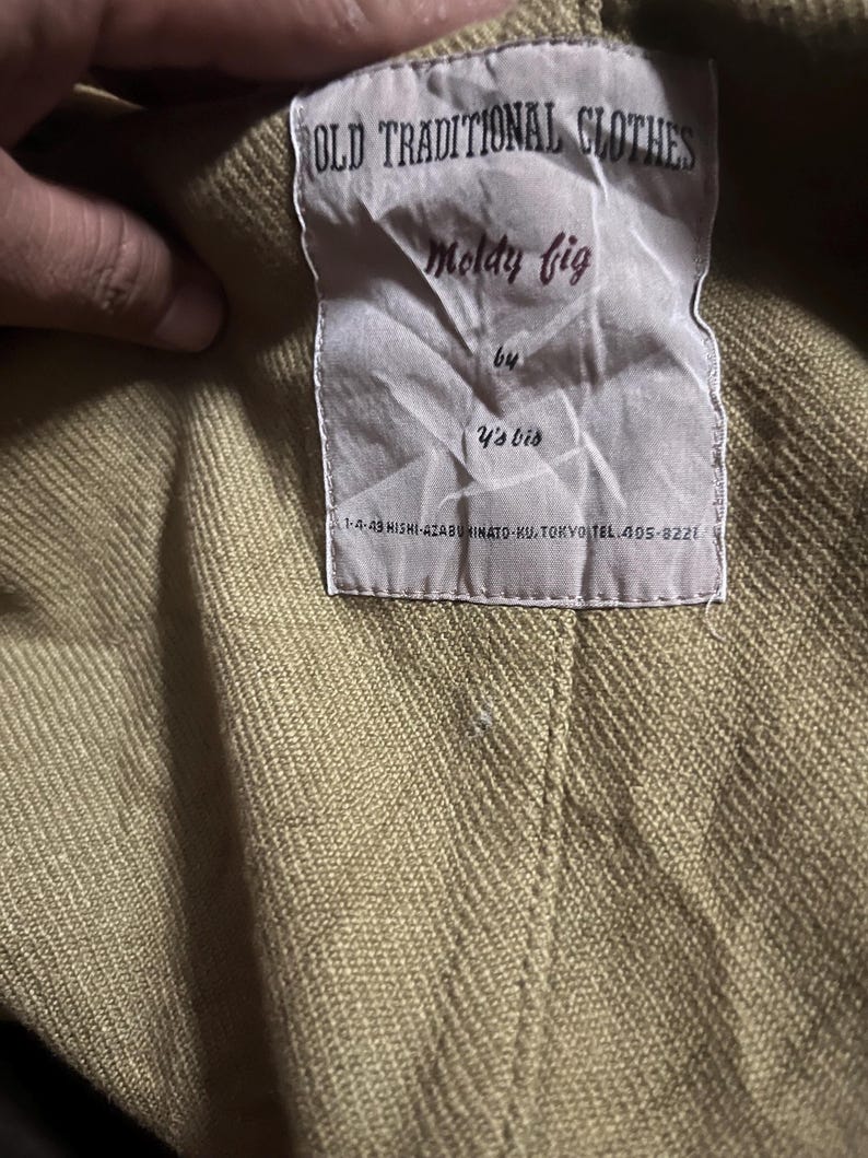 May include: A close-up of a clothing label. The label reads "OLD TRADITIONAL CLOTHES" at the top, with "Moldy fig" below. The label also includes "by Y's bis" and a Tokyo address. The garment is a light olive green colour.