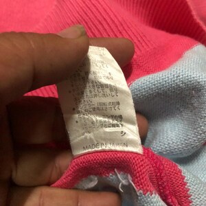May include: Close-up of a pink and light blue sweater. The sweater has a ribbed neckline and a tag that reads "MADE IN JAPAN". The tag has Japanese text. The sweater is made of a soft, knitted material.