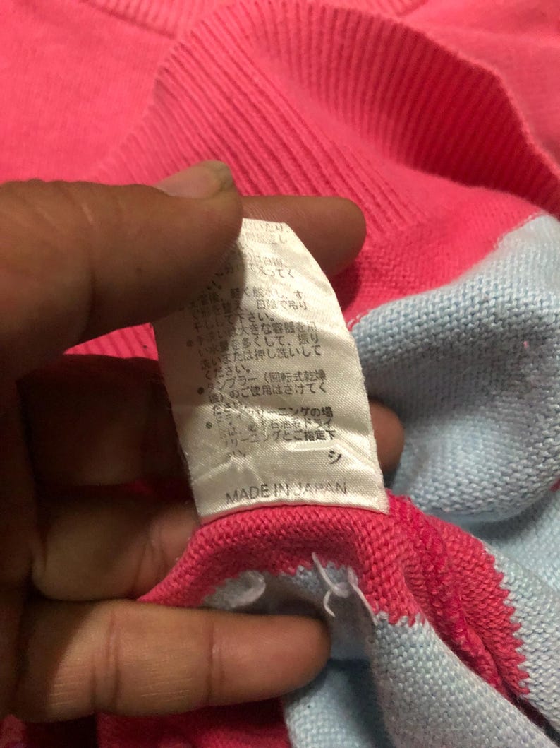 May include: Close-up of a pink and light blue sweater. The sweater has a ribbed neckline and a tag that reads "MADE IN JAPAN". The tag has Japanese text. The sweater is made of a soft, knitted material.