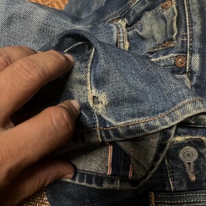 May include: Close-up of distressed blue denim jeans. The jeans show signs of wear and tear, with visible fading and a small hole near the pocket. The stitching is visible, and the fabric has a textured appearance. The jeans have metal rivets and buttons.
