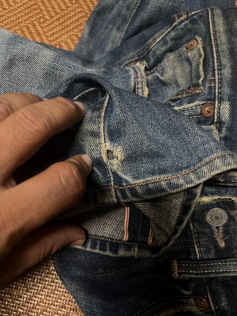 May include: Close-up of distressed blue denim jeans. The jeans show signs of wear and tear, with visible fading and a small hole near the pocket. The stitching is visible, and the fabric has a textured appearance. The jeans have metal rivets and buttons.