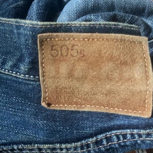 May include: Close-up of blue denim jeans with a brown leather patch. The patch has the number "505s" embossed on it. The jeans have visible stitching and a zipper.