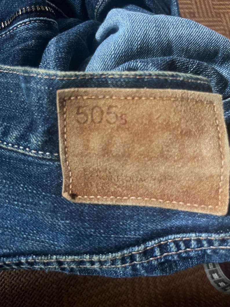 May include: Close-up of blue denim jeans with a brown leather patch. The patch has the number "505s" embossed on it. The jeans have visible stitching and a zipper.