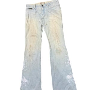 May include: Pale blue denim jeans with a faded wash and flared leg. The jeans have white embroidered floral patterns on the lower legs. They fasten with a button and zip.