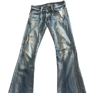 May include: Light blue denim jeans with a flared leg design. The jeans have a distressed look with a ripped section on the left leg. The jeans have a classic five-pocket design and a button and zipper closure.