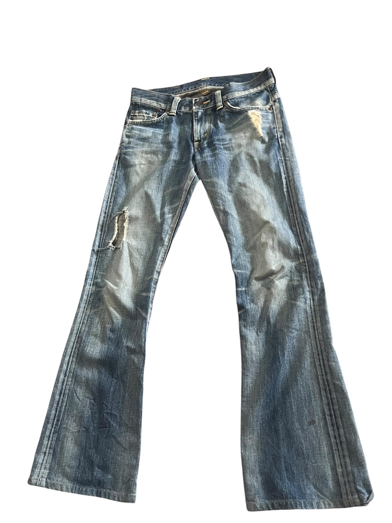 May include: Light blue denim jeans with a flared leg design. The jeans have a distressed look with a ripped section on the left leg. The jeans have a classic five-pocket design and a button and zipper closure.