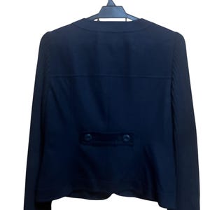 May include: A dark navy blue blazer hanging on a black hanger. The back of the blazer is visible, featuring a tailored design with a small, rectangular velvet detail and two buttons. The sleeves have a ribbed texture.