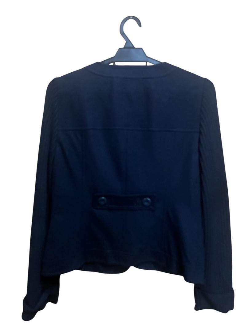 May include: A dark navy blue blazer hanging on a black hanger. The back of the blazer is visible, featuring a tailored design with a small, rectangular velvet detail and two buttons. The sleeves have a ribbed texture.