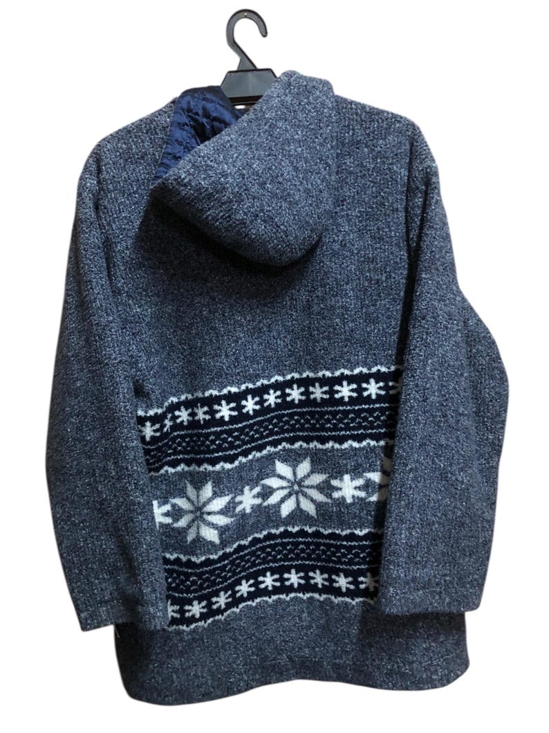 May include: A blue hooded sweater with a snowflake pattern on the lower back. The sweater is hanging on a black hanger. The snowflake design is white and navy blue.