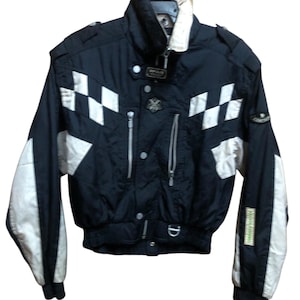 Peut inclure: Veste de ski noire et blanche avec un col montant et plusieurs fermetures éclair. La veste présente des touches blanches sur les manches et un motif à carreaux sur la poitrine. Un petit emblème est visible sur la poitrine.