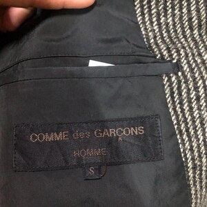 May include: Close-up of the interior of a jacket, featuring a black lining and a label that reads "COMME des GARCONS HOMME". The jacket's exterior is a black and white herringbone pattern. The label indicates a size "S".