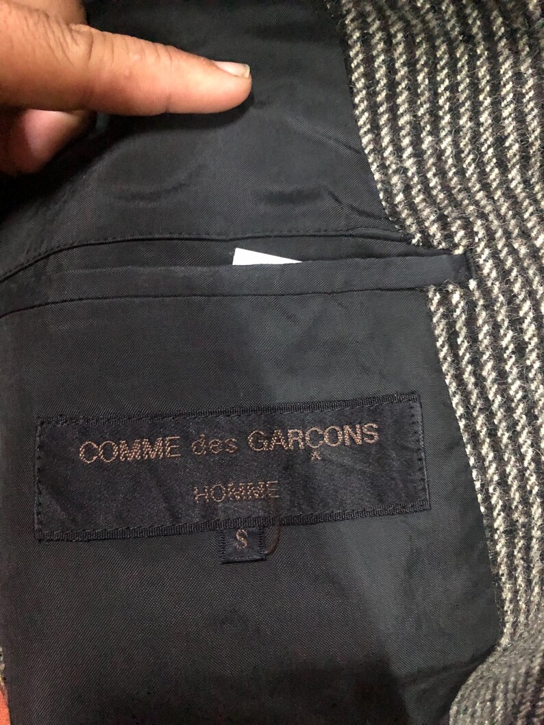 May include: Close-up of the interior of a jacket, featuring a black lining and a label that reads "COMME des GARCONS HOMME". The jacket's exterior is a black and white herringbone pattern. The label indicates a size "S".