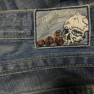 May include: Close-up of blue denim jeans with a True Religion patch. The patch features a skull and roses design with the brand name embroidered in white. The jeans have yellow stitching.