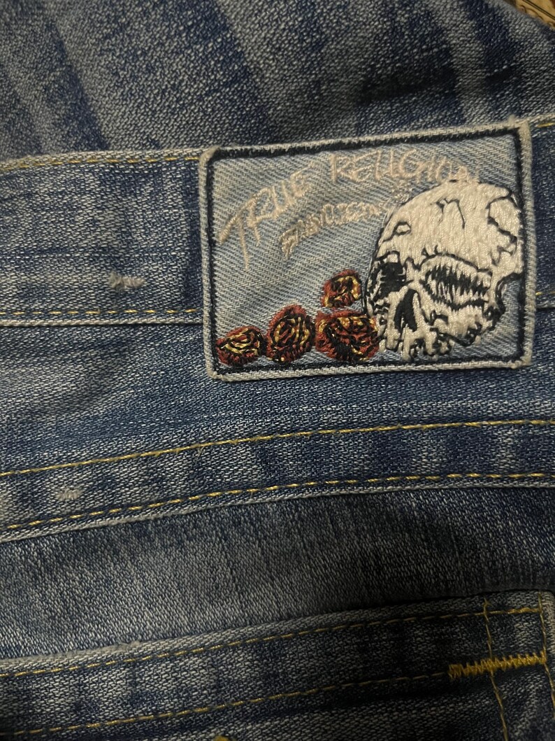 May include: Close-up of blue denim jeans with a True Religion patch. The patch features a skull and roses design with the brand name embroidered in white. The jeans have yellow stitching.