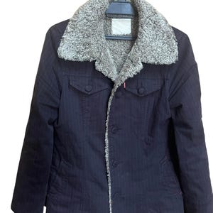 May include: A dark navy blue jacket with a faux fur collar and lining. The jacket has a pinstripe pattern, two chest pockets, and button closures. The jacket is hanging on a black hanger.
