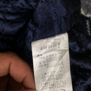 May include: Close-up of a navy blue quilted fabric with a white tag. The tag has Japanese text and numbers, including "ATN-98 39 W" and care symbols. The fabric appears to be part of a garment.