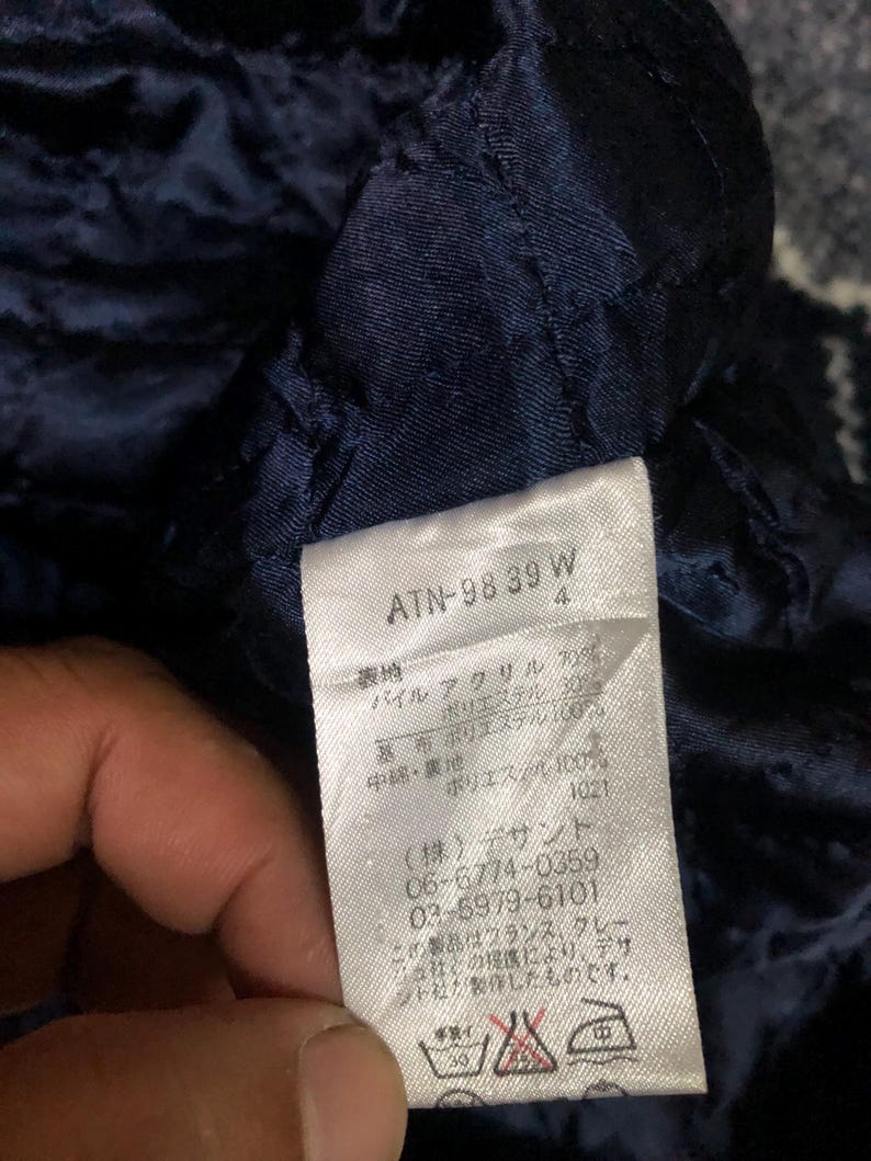 May include: Close-up of a navy blue quilted fabric with a white tag. The tag has Japanese text and numbers, including "ATN-98 39 W" and care symbols. The fabric appears to be part of a garment.
