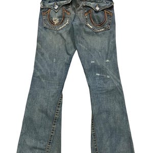 May include: Blue denim jeans featuring a distressed finish, two back flap pockets with button closures, and signature horseshoe stitching in orange and brown thread. A white True Religion brand patch is visible on the back waistband.