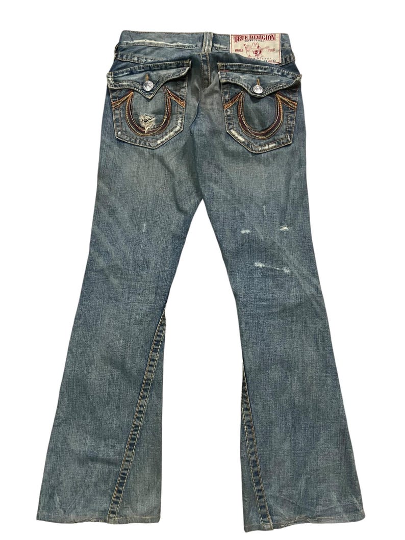 May include: Blue denim jeans featuring a distressed finish, two back flap pockets with button closures, and signature horseshoe stitching in orange and brown thread. A white True Religion brand patch is visible on the back waistband.