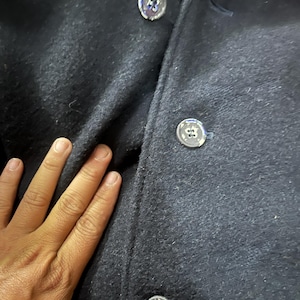 May include: Close-up of a navy blue wool coat. The coat features three silver buttons with a unique design. The fabric has a textured appearance. The image highlights the coat's details and material.