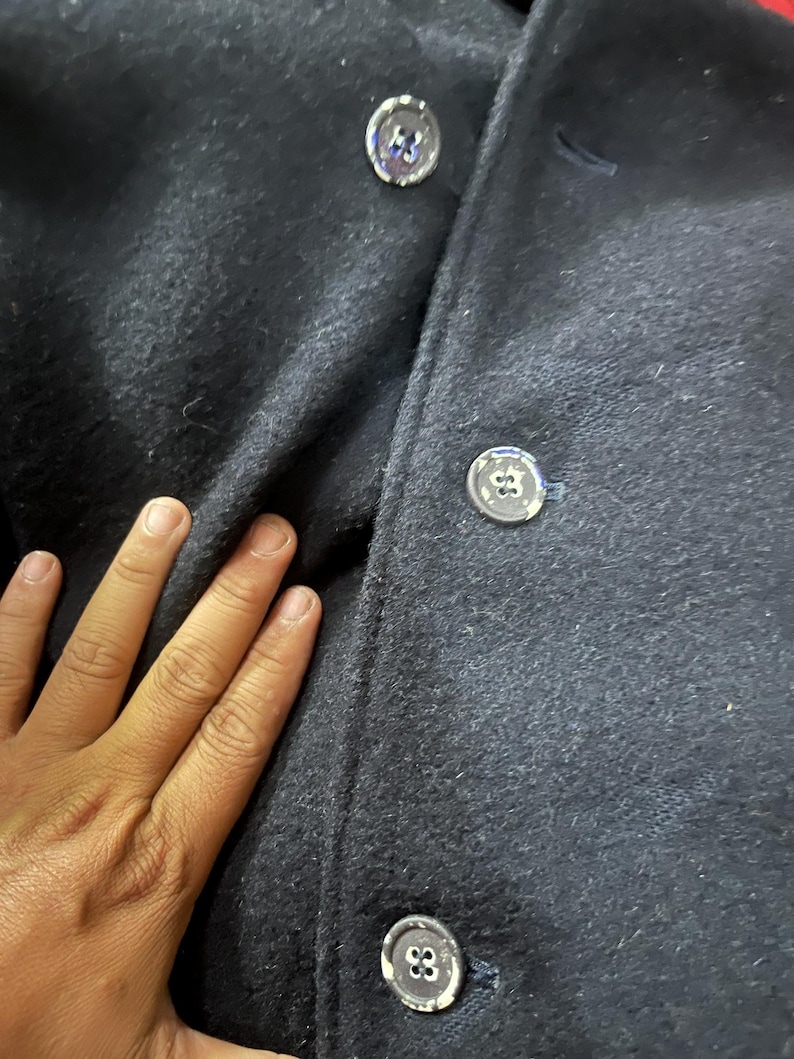 May include: Close-up of a navy blue wool coat. The coat features three silver buttons with a unique design. The fabric has a textured appearance. The image highlights the coat's details and material.