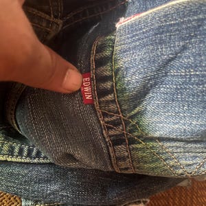 May include: Close-up of folded blue denim jeans. The red tag with the brand name "EDWIN" is visible. The jeans have visible stitching and a worn appearance.