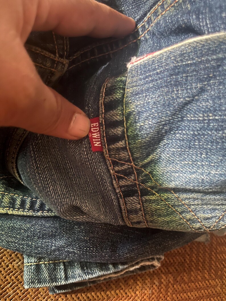 May include: Close-up of folded blue denim jeans. The red tag with the brand name "EDWIN" is visible. The jeans have visible stitching and a worn appearance.